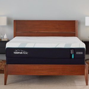 A wooden bed frame with a Tempur-Pedic TEMPUR-Adapt® 11" Medium Hybrid Mattress accents the bedroom. Two matching bedside tables with lamps and wicker baskets flank each side. The light-colored walls add a calming touch, while a window is partially visible, inviting natural light into the cozy space.