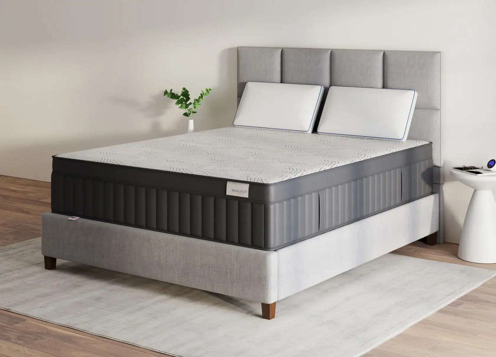 A modern bedroom features a queen-sized bed with a grey, upholstered frame and headboard. The bed is neatly made with a black and white NXT 2000 Firm Euro Top mattress and two pillows. A small white side table with decor is beside the bed, all atop a light-colored area rug.