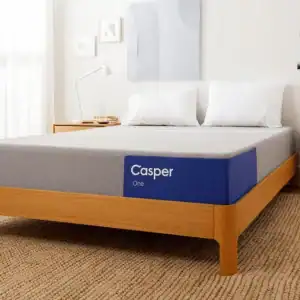 A minimalist bedroom featuring a Casper One mattress on a wooden bed frame. The room has white walls, light wooden flooring, a small bedside table, and some wall decorations.