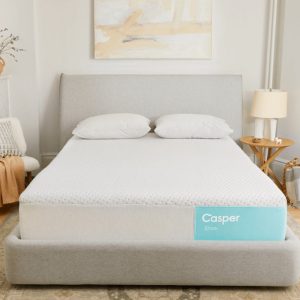 A modern bedroom featuring a neatly made bed with a gray headboard, topped with white bedding and pillows. A Snow mattress box is positioned at the foot of the bed. Cozy ambiance with a