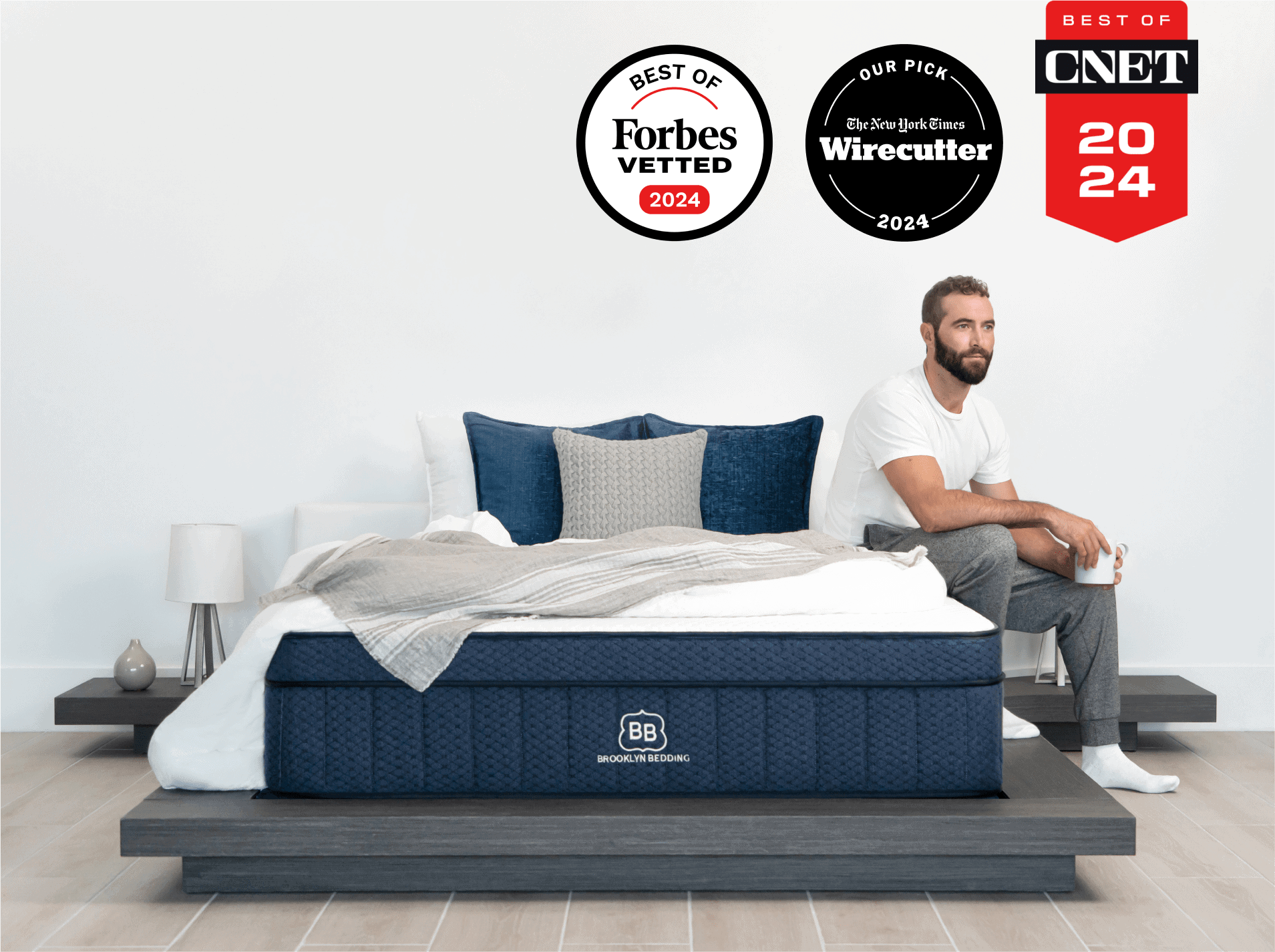 A man in a white shirt and gray pants sits on the edge of a modern bed with AURORA LUXE COOLING blue and white bedding. The bed is on a dark wooden platform. Above him are three awards: one from Forbes Vetted, one from Wirecutter, and one from CNET, all for 2024.