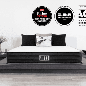 A modern bedroom features a neatly made PLANK FIRM bed, highlighting its black and white color scheme on a stylish platform frame. At the top, three award badges are displayed: Forbes Vetted Best Product 2024, Wired Recommends 2024, and ACA Endorsed.