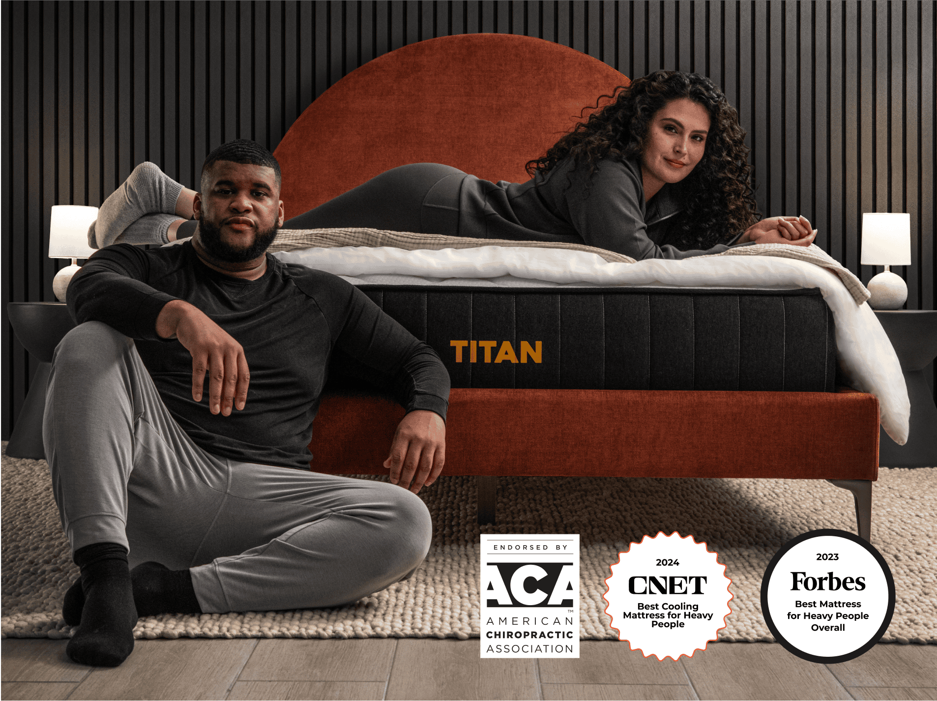 In a contemporary bedroom, a man sits on the floor while a woman relaxes on the Titan Plus mattress, showcasing its features. The display also highlights three awards from ACA, CNET, and Forbes for the Titan Plus's comfort and support.