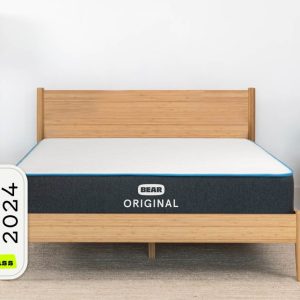 A minimalist bedroom showcases a wooden bed frame with the Bear Original mattress. Nearby, there is a dark blue upholstered chair decorated with a cushion that features an American flag pattern. To the left stands a floor lamp, and prominently displayed is the "Best in Class 2024" award from The Strategist.