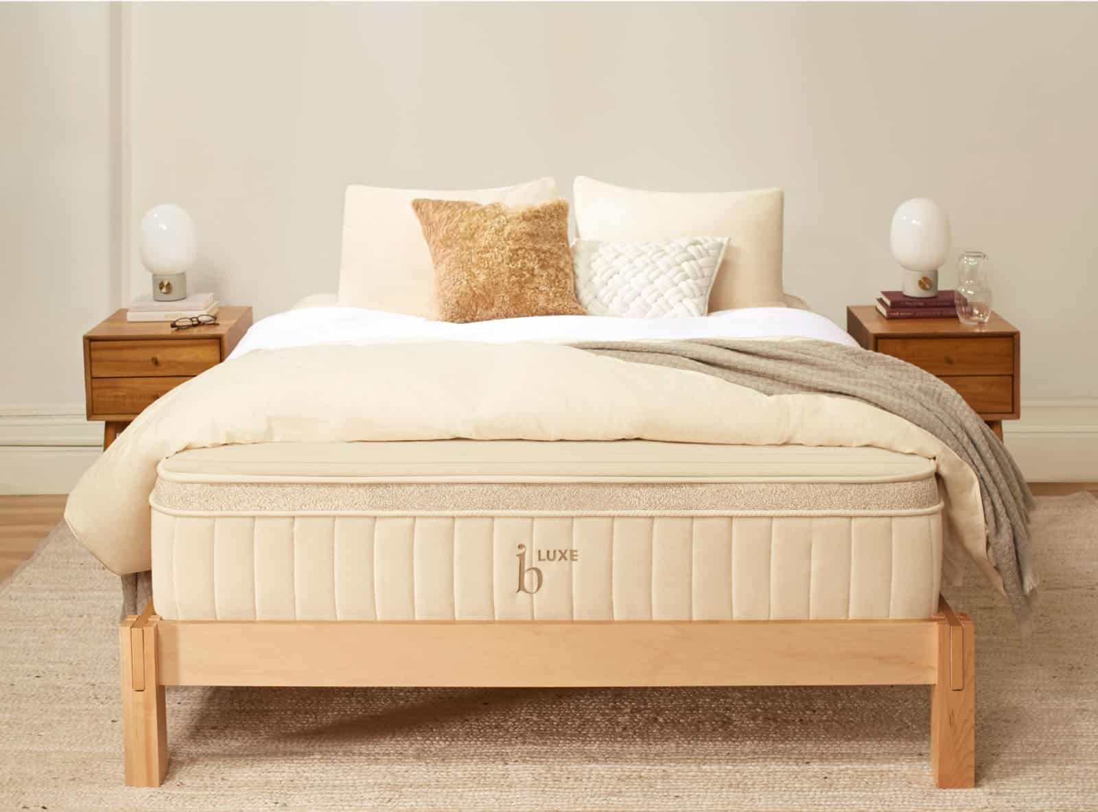 A neatly made bed with a luxe Birch Luxe Natural Mattress and a light-colored wooden frame is centered in the room. The bed is topped with a variety of white and beige pillows, a textured throw, and surrounded by birch nightstands with lamps and minimalist decor.
