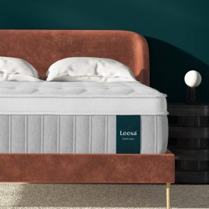 A stylish bedroom showcases a soft orange plush velvet bed frame, outfitted with the luxurious Sapira Chill Hybrid Mattress. Next to the bed stands a sleek black bedside table adorned with a small, spherical white lamp. The room is highlighted by dark teal walls and complemented by a beige carpet.