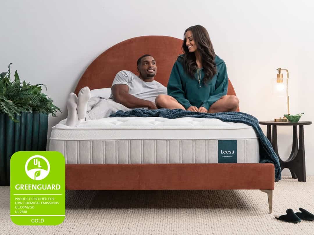 A man lies on a Sapira Chill Hybrid Mattress while a woman sits next to him. The bed is positioned in front of a brown, semi-circular headboard, with a plant and lamp on either side. A green "GREENGUARD" certification badge is visible in the bottom-left corner.