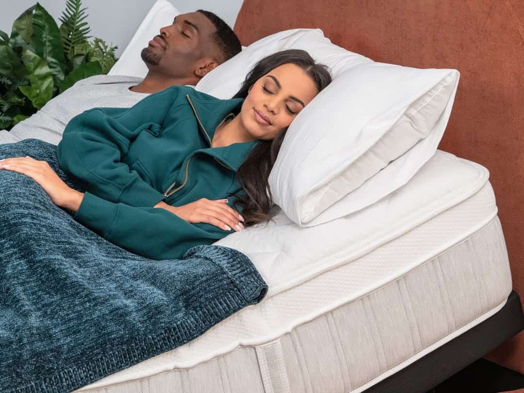 A man and woman are lying on a bed, both peacefully sleeping on a Sapira Chill Hybrid Mattress. The woman, wearing a teal zip-up jacket, is covered with a blue blanket, while the man in a light gray shirt enjoys the cool ambiance. Both rest on white pillows, and there are green plants in the background.