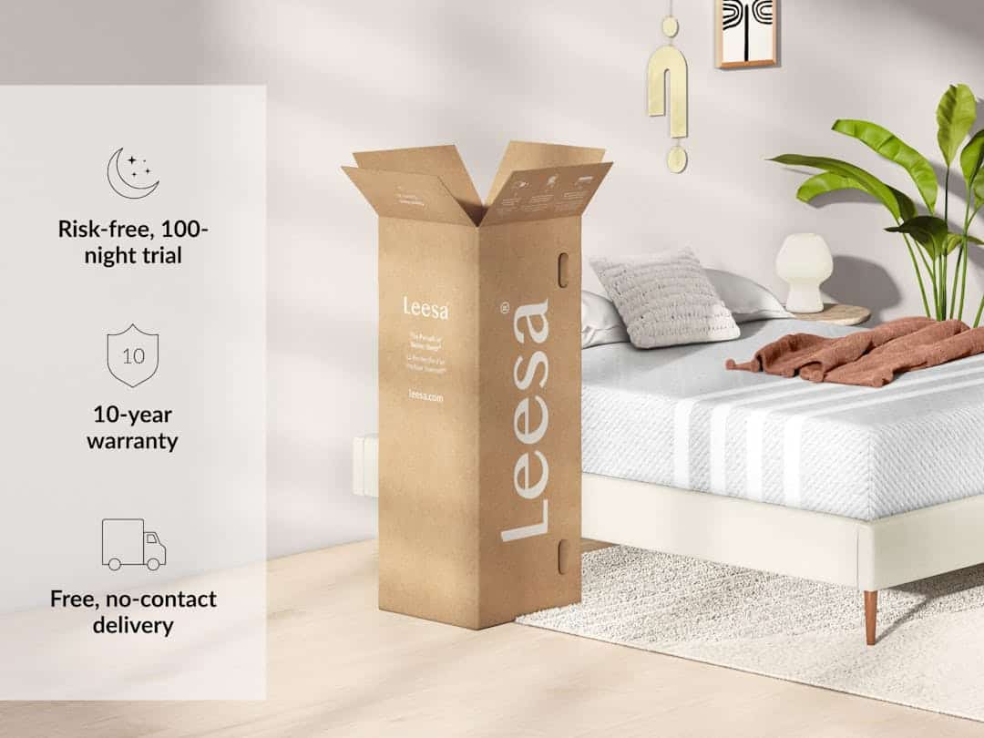 A bedroom scene showcasing a bed with a white mattress and pillows, beside an open cardboard box labeled "Leesa Original Hybrid Mattress." The image highlights three features: a risk-free, 100-night trial; a 10-year warranty; and free, no-contact delivery. The room has modern, minimalistic decor.