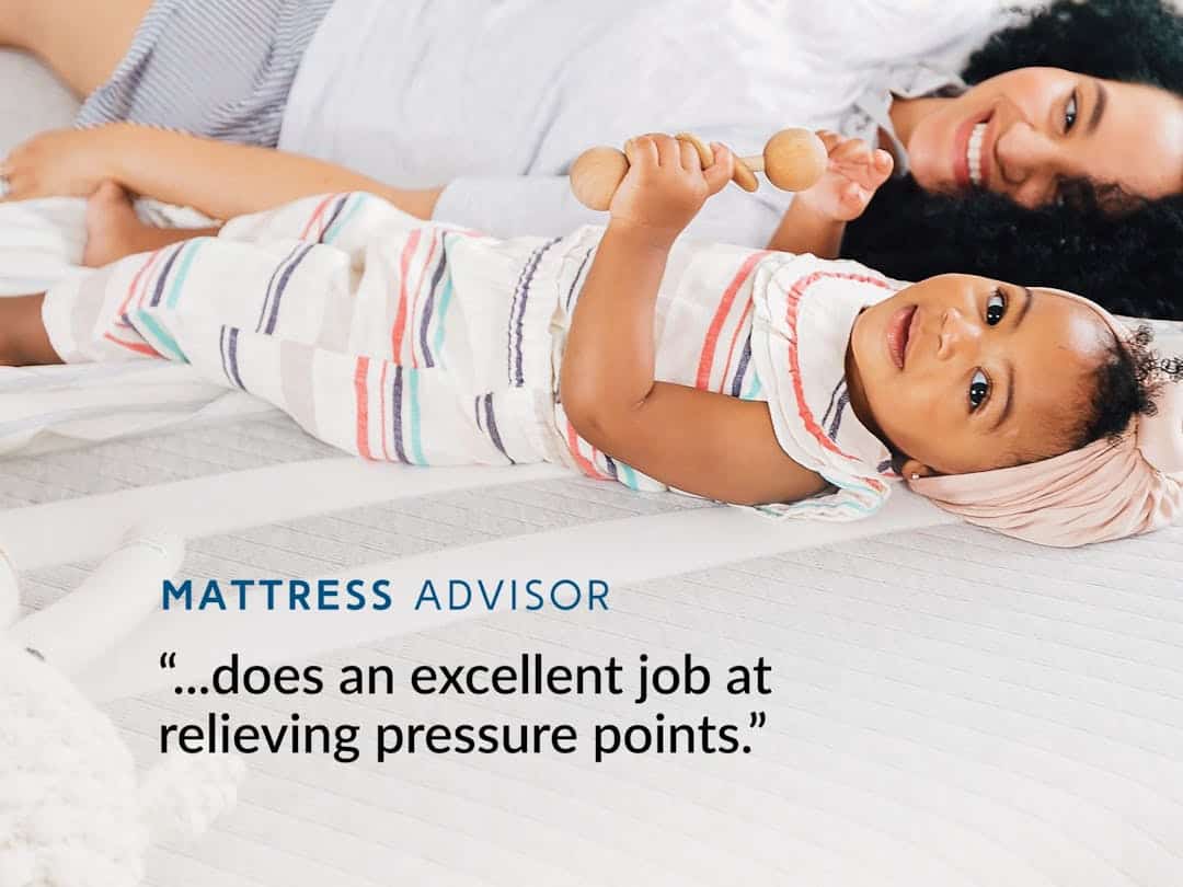 A mother and her baby lie comfortably on an Original Hybrid Mattress. The baby, dressed in a striped outfit and headband, holds maracas while the mother smiles in the background. The text reads "MATTRESS ADVISOR: The Original Hybrid does an excellent job at relieving pressure points.