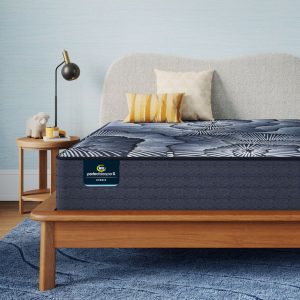 A modern bedroom with a neatly made bed featuring a Serta Perfect Sleeper X Hybrid Mattress: Aspen Firm adorned with a grey and black patterned cover. The bed has a light grey upholstered headboard. A round wooden nightstand beside the bed holds a black lamp and small decor items, including a plush sheep. The room has a textured light blue wall and a blue rug.