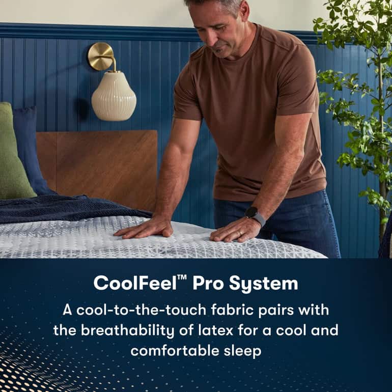 A man in a brown shirt is pressing down on a Serta iComfortPro Mattress: Chelsea Medium with his hands. Behind him, a plant, bed with green and navy pillows, and a wall-mounted light can be seen. The text on the image reads, "CoolFeel™ Pro System: A cool-to-the-touch fabric pairs with the breathability of latex for a cool and comfortable sleep.