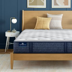 A neatly made bed with a light wooden frame in a stylish room showcases the ultimate comfort of the Serta iComfortPro Mattress: Chelsea Medium. Adorned with two pillows—one plain white and one patterned yellow—a round white nightstand with a lamp and decor is positioned to the left of this cozy Chelsea Medium bed.