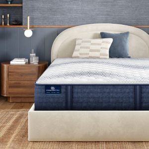 A modern bedroom with a beige upholstered bed frame and a Serta iComfortPro Mattress: Kingston Firm. The bed is adorned with a mix of patterned and solid pillows. A wooden nightstand with a drawer holds a lamp, a book, and a decorative item. A textured rug covers the floor.