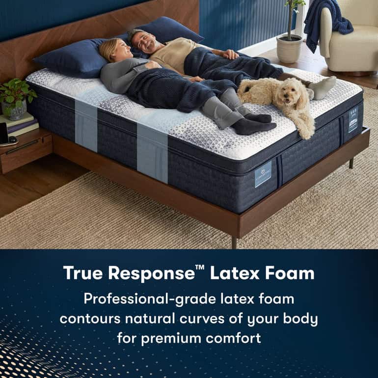 A couple lies comfortably on a bed with a dog at their feet. The Serta iComfortPro Mattress: Chelsea Medium, featuring "True Response™ Latex Foam," contours to the natural curves of the body for premium comfort. A plant and nightstand are on either side of the bed.