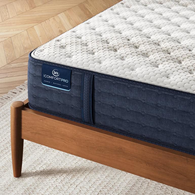 A close-up of a bed featuring the Chelsea Medium Serta iComfortPro mattress with a white, patterned surface labeled "Serta iComfort Pro" on a dark blue base. The mattress is supported by a wooden bed frame with angled legs, positioned atop a light-colored textured rug over a wooden floor arranged in a herringbone pattern.