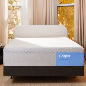A minimalist bedroom featuring the Casper Dream Max: Hybrid Mattress on a bed frame with a beige headboard. The room includes light-colored curtains, large windows showcasing autumn foliage, a black floor lamp, a bedside table with decorations, and a wooden dresser.
