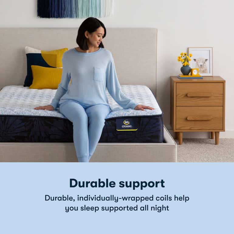 A woman sits on the edge of a bed wearing a light blue sweater and matching pants. The bed features a Serta Classic Mattress with blue and white bedding. A wooden nightstand with books, flowers, and a lamp stands beside the bed. Text reads “Durable support.”
