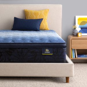 A neatly made bed featuring a Serta Classic Mattress: Brecon Plush Pillow Top with two pillows, one dark blue and one yellow with a wavy pattern. The bed has a beige upholstered headboard. A wooden nightstand with a small vase, a framed abstract picture, and a yellow-tinted alarm clock is beside it.