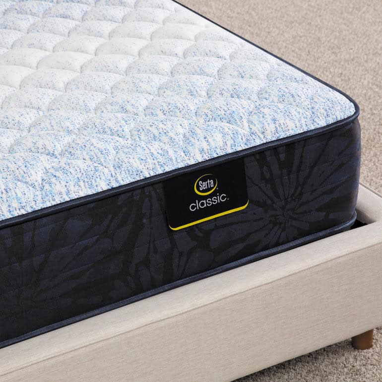 A close-up of the corner of a bed featuring a Serta Classic Mattress with a textured surface. The mattress, labeled "Serta Classic," rests on a beige bed frame. The floor underneath is covered with light-colored carpet.