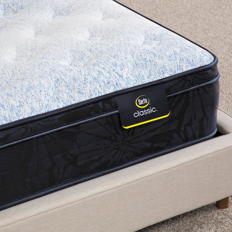 A close-up of the Serta Classic Mattress: Dorian Plush Euro Top showcasing its white and blue speckled top layer and black, patterned side. The mattress tag reads "Dorian Plush." It is placed on a light beige bed frame, against a backdrop of beige, carpeted floor.
