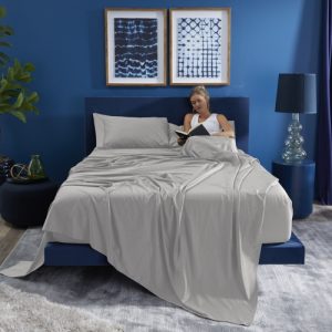 A person relaxes in bed, engrossed in a book and draped in the light gray Basic Sheet Set. The room features blue walls, a matching blue bed frame, and a dark blue nightstand that holds a bottle and plant. A clear lamp with a dark blue shade casts light on two framed abstract artworks positioned above the bed.