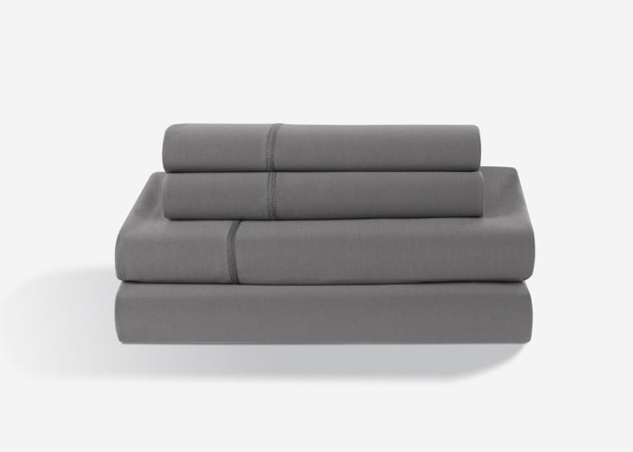 A neatly folded Dri-Tec Sheet Set in grey stands arranged in a stack against a plain white background. This set comprises two pillowcases, a fitted sheet, and a flat sheet, all in matching grey fabric. The top pillowcase features a subtle stitched line detail.