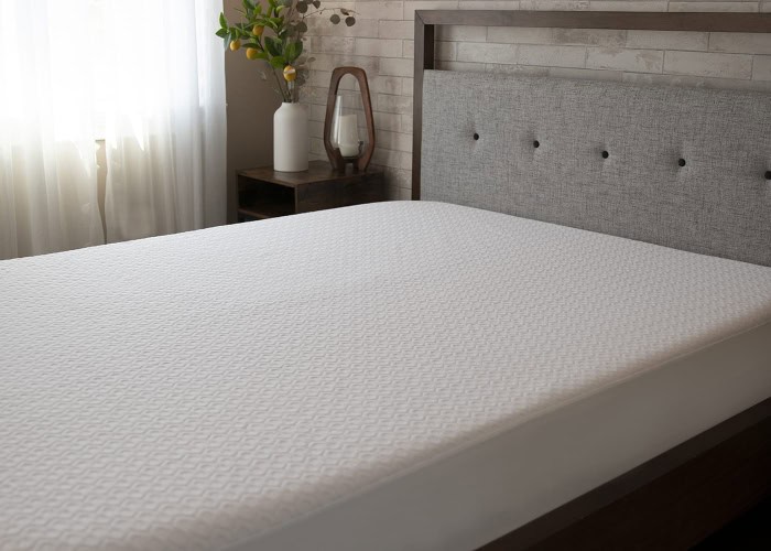 A neatly made bed with a white Dri-Tec Mattress Protector takes center stage in a well-lit bedroom. The bed features a button-tufted grey headboard, while a wooden side table in the background holds a white vase with green branches and a small decorative mirror.