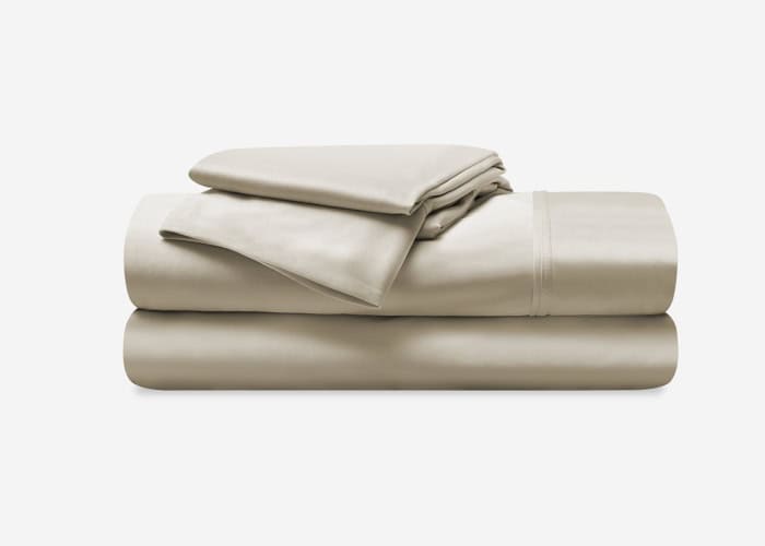 Against a plain white background, a neatly folded Dri-Tec Sheet Set in beige is arranged in a stack, featuring a fitted sheet, flat sheet, and pillowcases. The sheets exhibit a smooth, silky texture.
