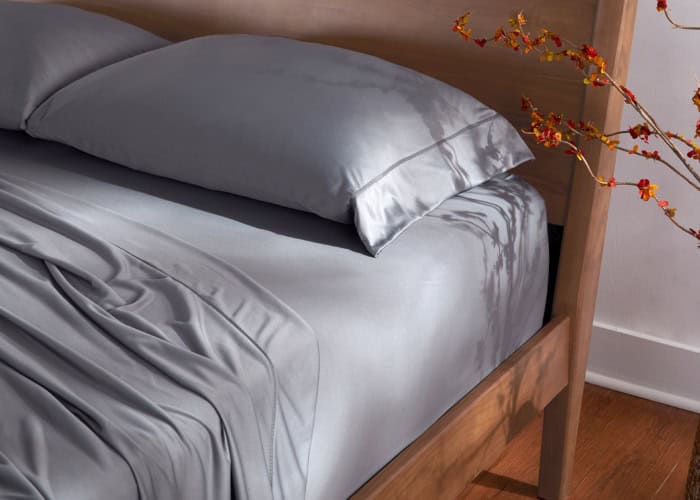 A neatly made bed with a Dri-Tec Sheet Set in light gray and matching pillows rests within a wooden frame, bathed in soft, natural light. At the edge, a few orange and red flowers on branches cast gentle shadows on the bed, creating a serene and cozy atmosphere.