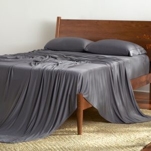 A wooden bed with a headboard, adorned with a Dri-Tec Sheet Set in gray that elegantly drapes over the sides, includes two matching pillowcases. The bed is positioned on a woven rug within a room featuring a hardwood floor. Adjacent to the bed, against a plain wall, stands a tall woven vase filled with branches.