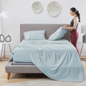 A woman is making a bed with a light blue Hyper – Linen Sheet Set in a well-lit bedroom. She is holding a pillow and adjusting the pillowcase. The room has modern decor, including white and grey accents, decorative wall pieces, and a small lamp on a bedside table.