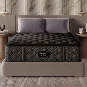 A modern bedroom features a queen-sized bed with a Beautyrest Black® Series Three 15.25" Medium Mattress and a beige upholstered headboard. The bed is flanked by two round wooden nightstands, each holding a lamp and decorative items. Neutral tones and minimalistic decor create a serene atmosphere.