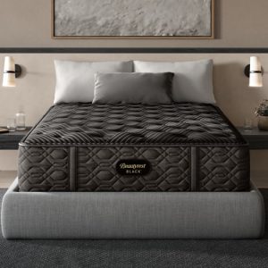 A modern bedroom features a double bed with a Beautyrest Black® Series One 13.5" Extra Firm Mattress. The bed has gray upholstered sides, and the room includes two floating nightstands with wall-mounted lights. Decorative items like books and plants are placed on the nightstands.