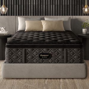 A modern bedroom featuring a plush, black "Beautyrest Black® Series Three 16.25" Medium Pillow Top Mattress" on a gray platform bed. The bed is flanked by two minimalist black nightstands, each adorned with hanging pendant lights, books, and a small potted plant. Neutral-toned pillows contrast the bed's dark aesthetic.