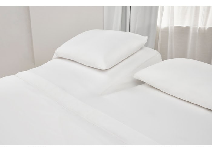 A neatly made bed featuring the premium Hyper-Cotton Sheet Set, with its crisp white linens perfectly arranged, including two pillows and a blanket from the collection. The room exudes a bright and clean appearance, accentuated by white curtains that partially cover a window in the background. The overall ambiance feels calm and inviting.