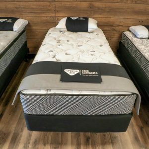 Three neatly arranged beds with Spring Air 200 Plush mattresses are on display in a store. Each bed is accompanied by a pillow and draped with a black cloth featuring text, all set against the backdrop of a wooden floor. A wood-paneled wall behind them enhances the warm atmosphere.