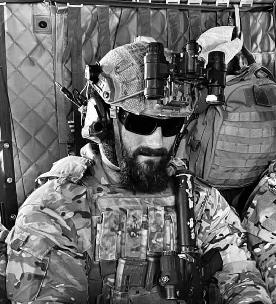 A soldier in tactical gear sits inside a vehicle. He is wearing a helmet with night vision goggles, sunglasses, and a radio headset. The interior is lined with quilted material. The image is in black and white.