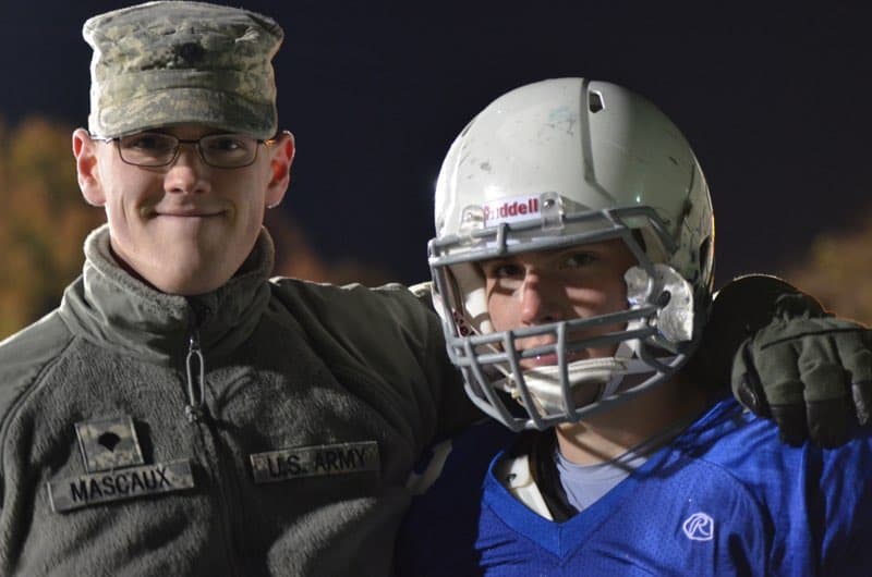 A U.S. Army officer wearing a uniform and cap stands with his arm around a football player in a helmet and blue jersey. Both are smiling at the camera. They appear to be outdoors, possibly at a sports field.