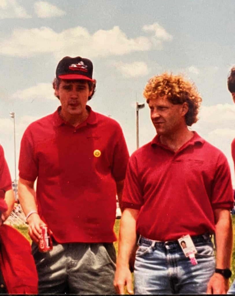 Two people wearing red shirts and standing outdoors. One person is holding a can of soda and wearing a baseball cap. Both are under a partly cloudy sky, and there's a grassy area in the background.