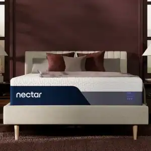 A stylish bedroom with a modern bed featuring a Nectar Premier Memory Foam Mattress Copy. The bed is adorned with burgundy and gray pillows and a quilt, flanked by wooden side tables with lamps. The walls are maroon, and light filters through the windows.