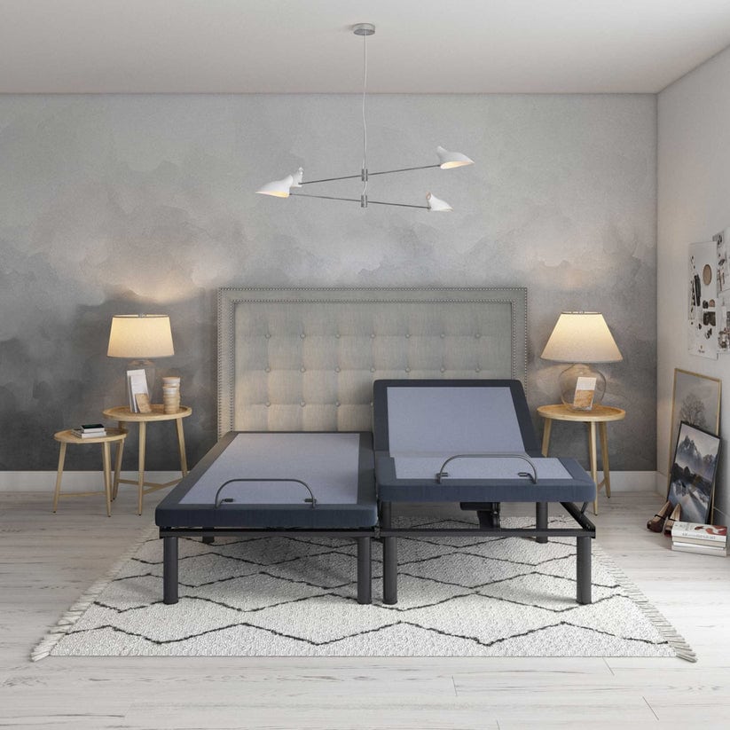 A modern bedroom with a Bed Tech 6500 Adjustable Base split into two sections, each slightly elevated. It features a gray tufted headboard, two round wooden side tables with lamps, and a neutral-toned rug on light wood flooring, embodying innovation and comfort.