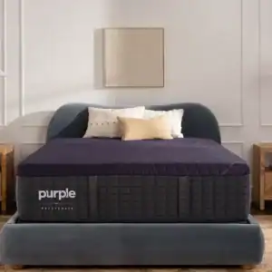 A modern bedroom showcases the Purple Rejuvenate Mattress on a plush gray bed frame, complemented by neutral pillows and nightstands, all set against a white paneled wall adorned with abstract art.