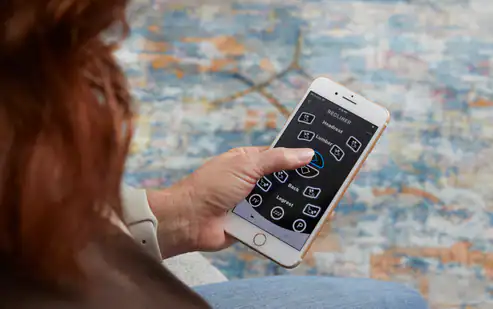 A person with long brown hair uses a smartphone with large app icons while relaxing in a Zecliner Model 2 Petite — Power Sleep Chair, against a blurred, colorful background.