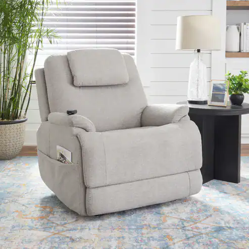 A light gray Zecliner Model 2 Petite Power Lift Recliner with headrest sits on a patterned rug beside a round black side table holding a lamp, glass vase, and book. A potted plant and window blinds are in the background.