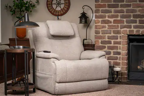 A beige Zecliner Model 1 Power Sleep Chair with a headrest is placed next to a round side table and lamp in a cozy living room featuring a brick fireplace, clock, and wall-mounted lantern.