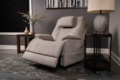 A Zecliner Model 2 Petite Power Lift Recliner in light gray with a headrest sits on a patterned rug, placed between a side table with a lamp and another table with a vase in a modern, softly lit living room.
