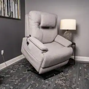 A Zecliner Model 2 Petite Power Lift Recliner in light gray sits partially raised in a modern living room, with its remote on the armrest. The space includes a table, lamp, plant, rug, and abstract wall art.
