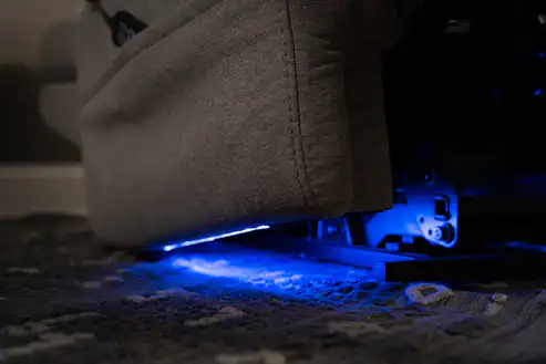 Close-up of the bottom edge of the Model 2 Plus Power Lift Recliner in gray, featuring blue LED lights that illuminate the floor and highlight part of the chair’s metal mechanism.