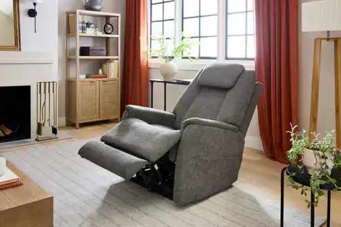 An Umber Zecliner Model 3+ Power Sleep Chair is extended in a cozy, well-lit living room with large windows, orange curtains, plants, a bookshelf, and a fireplace.